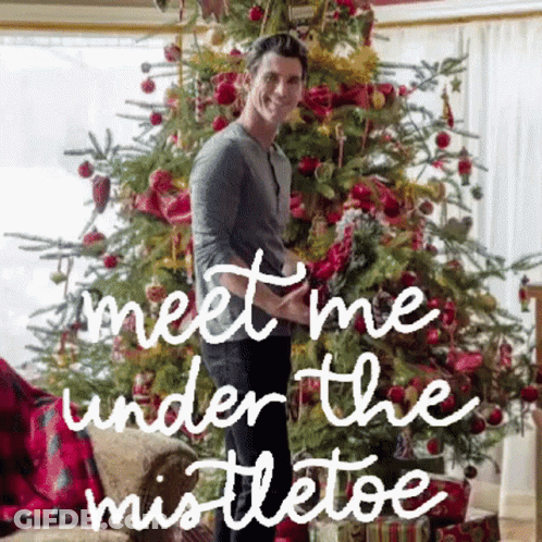 A Man Is Standing In Front Of A Christmas Tree With The Words `` Meet Me Under The Mistletoe '' . GIF