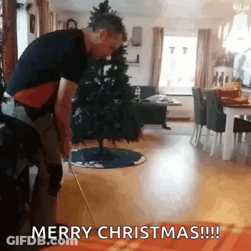 A Man Is Standing In Front Of A Christmas Tree With The Words Merry Christmas Written On The Bottom GIF