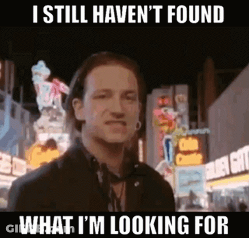 A Man Is Standing In Front Of A City Street At Night With A Sign That Says I Still Haven 'T Found GIF