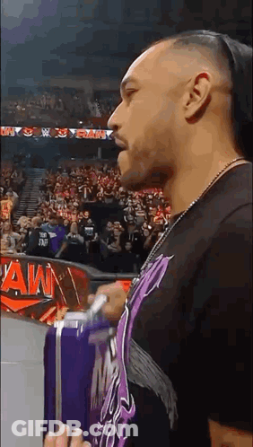 A Man Is Standing In Front Of A Crowd In A Stadium Holding A Purple Suitcase . GIF