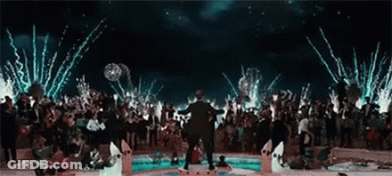 A Man Is Standing In Front Of A Crowd Of People At A Party With Fireworks Going Off In The Background . GIF