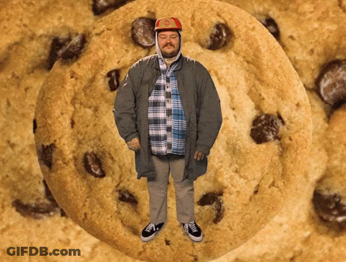 A Man Is Standing In Front Of A Large Cookie With Chocolate Chips GIF