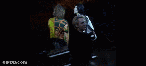 A Man Is Standing In Front Of A Piano On A Stage While A Woman Stands Behind Him . GIF