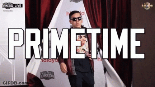 A Man Is Standing In Front Of A Red Curtain With The Words Primetime Written Above Him GIF