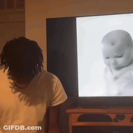 A Man Is Standing In Front Of A Television With A Baby On The Screen GIF