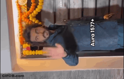 A Man Is Standing In Front Of A Wall With Flowers And A Sign That Says Aura 1577+ On It GIF