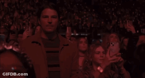 A Man Is Standing In The Middle Of A Crowd Of People Taking Pictures Of A Concert . GIF