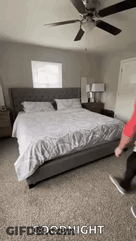 A Man Is Standing Next To A Bed In A Bedroom With A Ceiling Fan And A Window . GIF