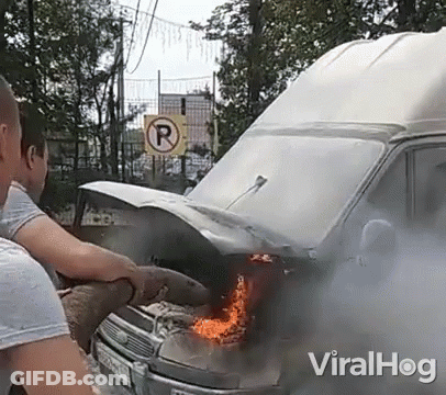 A Man Is Standing Next To A Burning Van With Smoke Coming Out Of It . GIF