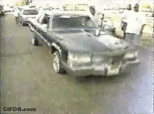 A Man Is Standing Next To A Car That Is Driving Down A Street . GIF