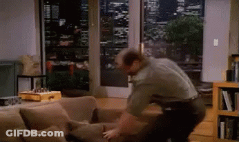 A Man Is Standing Next To A Couch In A Living Room Looking Out A Window GIF