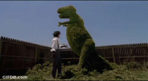 A Man Is Standing Next To A Dinosaur Made Of Bushes . GIF