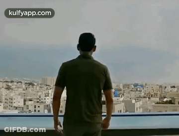 A Man Is Standing On A Balcony Overlooking A City And A Swimming Pool . GIF