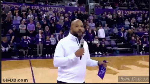 A Man Is Standing On A Basketball Court Holding A Microphone In Front Of A Crowd Of People . GIF