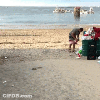 A Man Is Standing On A Beach Next To A Green Barrel That Says ' Coca Cola ' On It GIF