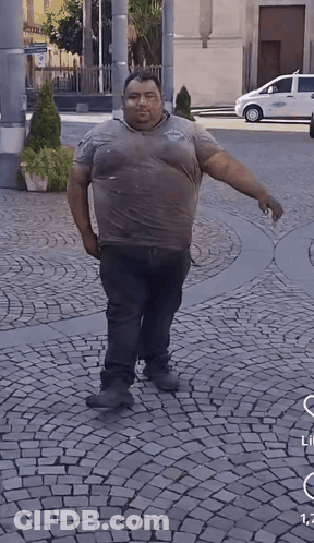 A Man Is Standing On A Cobblestone Street With A White Car Parked Behind Him GIF