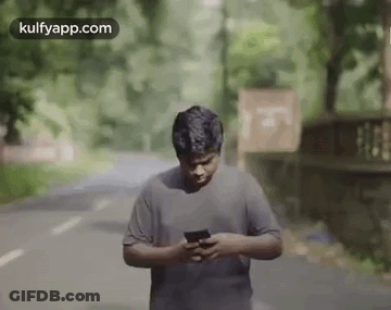 A Man Is Standing On A Road Looking At His Phone . GIF