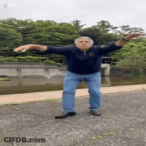 A Man Is Standing On A Sidewalk With His Arms Outstretched In Front Of A River . GIF