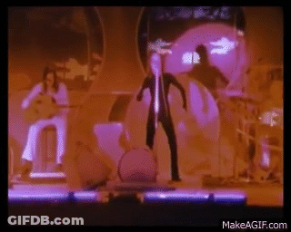 A Man Is Standing On A Stage In Front Of A Drum Set While A Woman Plays A Guitar . GIF