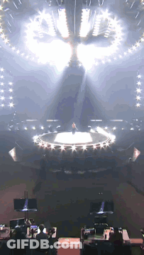 A Man Is Standing On A Stage With A Circle Of Lights Around Him GIF