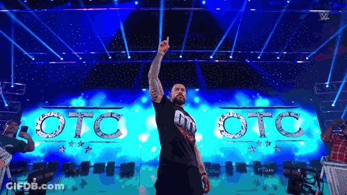 A Man Is Standing On A Stage With His Hand In The Air In Front Of A Sign That Says Oto Oto GIF