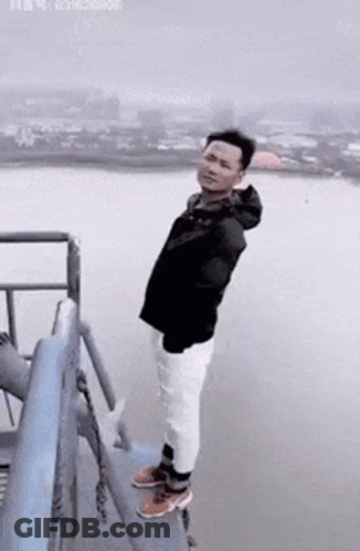 A Man Is Standing On The Edge Of A Bridge Overlooking A Body Of Water . GIF
