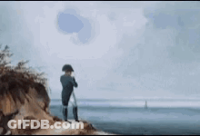 A Man Is Standing On Top Of A Cliff Overlooking The Ocean Looking Through Binoculars . GIF