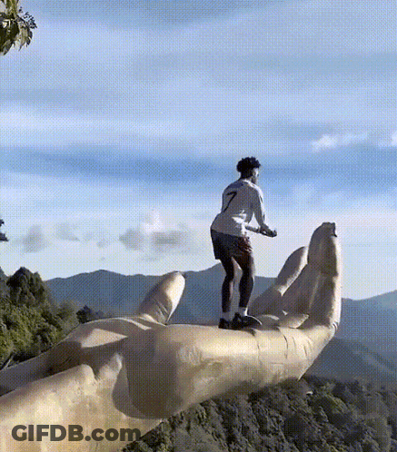 A Man Is Standing On Top Of A Giant Hand With Mountains In The Background GIF