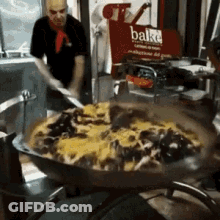 A Man Is Stirring A Large Pan Of Food In Front Of A Sign That Says Bake GIF