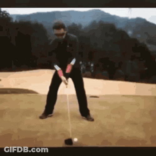 A Man Is Swinging A Golf Club At A Ball On A Golf Course . GIF
