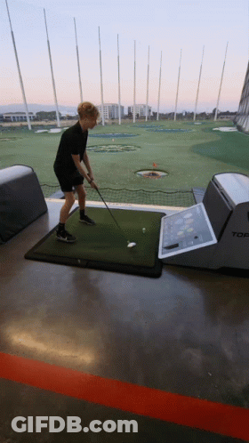 A Man Is Swinging A Golf Club At A Golf Ball On A Mat That Says Top GIF