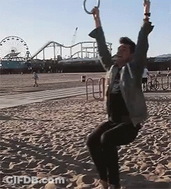 A Man Is Swinging On A Rope Swing On A Beach With A Roller Coaster In The Background GIF