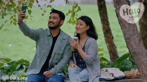 A Man Is Taking A Selfie With A Woman While They Sit Under A Tree With A Hello Again Sign Behind Them GIF