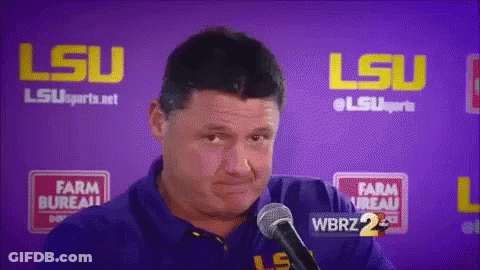 A Man Is Talking Into A Microphone In Front Of A Purple Wall With Lsu Written On It GIF