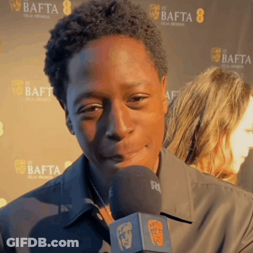 A Man Is Talking Into A Microphone In Front Of A Wall That Says Bafta GIF