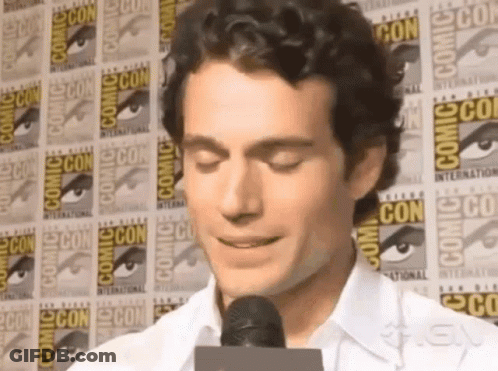 A Man Is Talking Into A Microphone In Front Of A Wall That Says Comic Con GIF