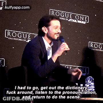 A Man Is Talking Into A Microphone While Sitting At A Table In Front Of A Sign That Says Rogue One . GIF