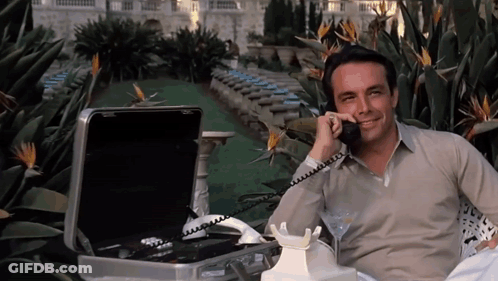 A Man Is Talking On A Phone Next To A Suitcase GIF