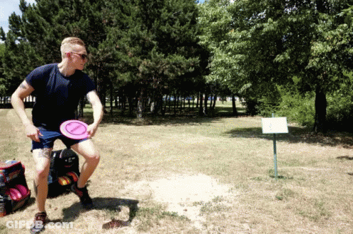 A Man Is Throwing A Frisbee In A Field With Trees In The Background And A Sign That Says " A " GIF
