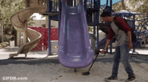 A Man Is Using A Metal Detector In A Playground In Front Of A Slide GIF