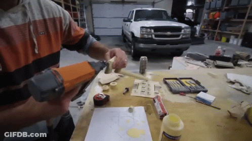 A Man Is Using A Nail Gun On A Piece Of Wood In Front Of A Truck GIF