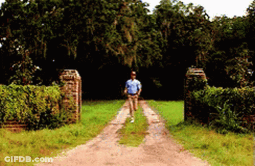 A Man Is Walking Down A Dirt Road In A Field GIF