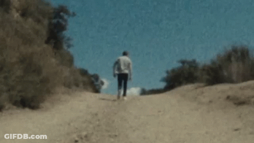 A Man Is Walking Down A Dirt Road In The Woods . GIF