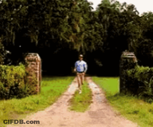 A Man Is Walking Down A Dirt Road Surrounded By Trees GIF