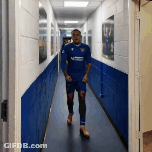 A Man Is Walking Down A Hallway Wearing A Blue Shirt That Says Mandy GIF
