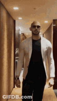 A Man Is Walking Down A Hallway Wearing Sunglasses And A White Jacket . GIF