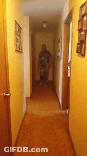 A Man Is Walking Down A Hallway With Pictures On The Walls GIF