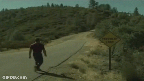 A Man Is Walking Down A Road Next To A Sign That Says Pavement Ends GIF