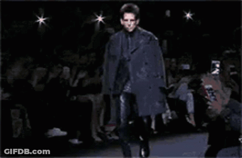 A Man Is Walking Down A Runway Wearing A Black Coat GIF