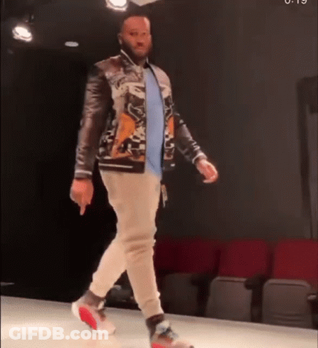 A Man Is Walking Down A Runway Wearing A Jacket And Sweatpants . GIF
